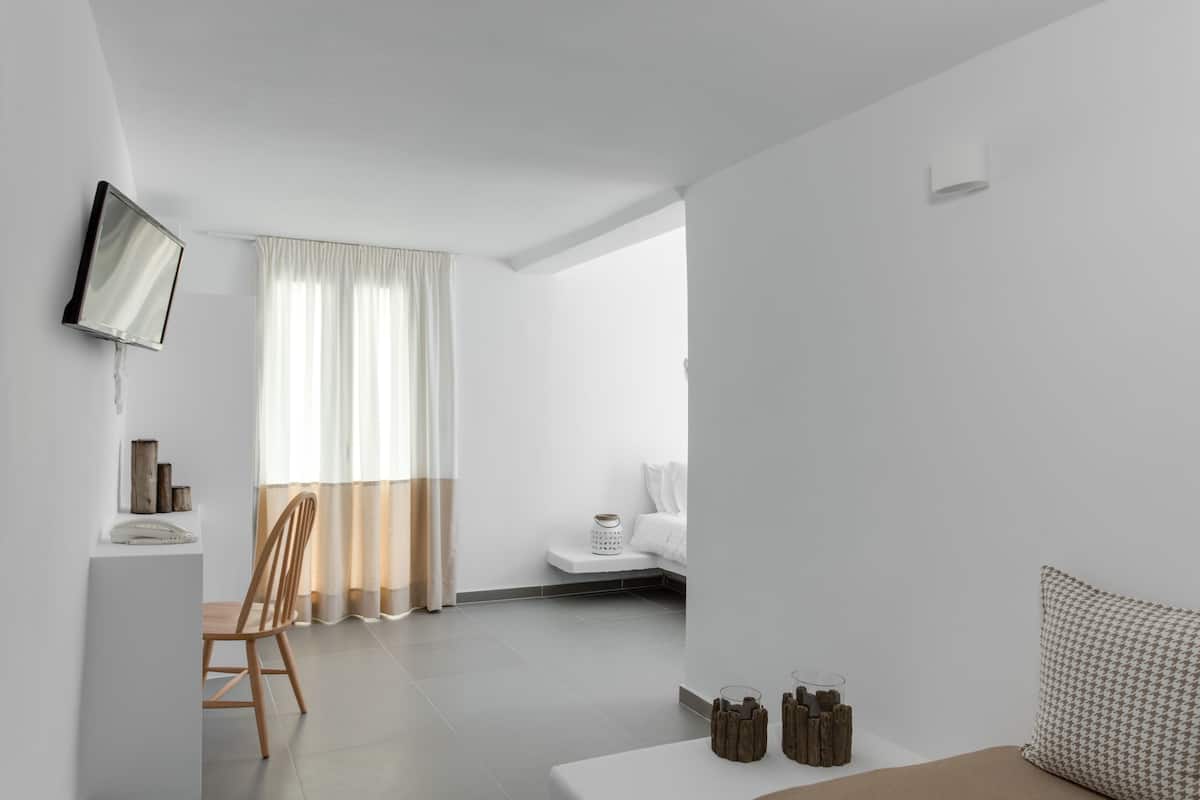 A modern, minimalist room with a flat-screen TV mounted on the wall, a wooden chair, a small table with decorative items, a bed with white bedding, and a large window with sheer curtains.