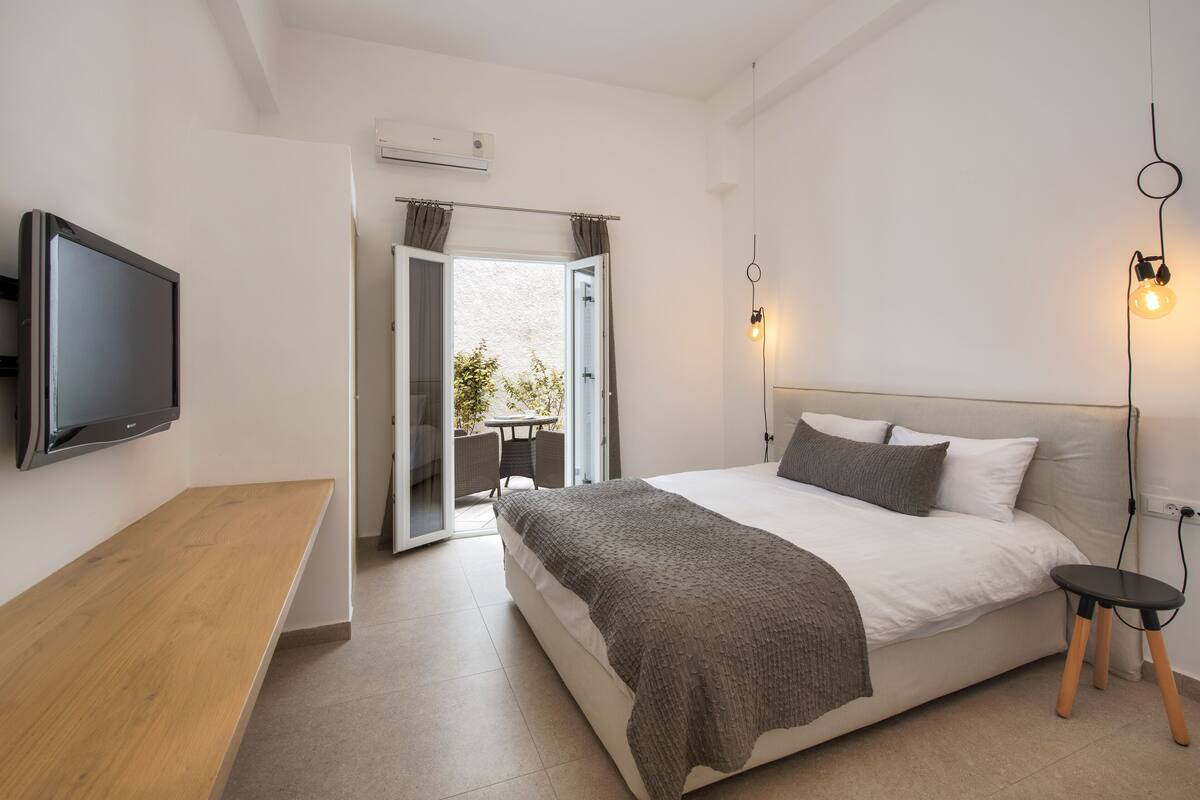 A bedroom with a bed, a TV mounted on the wall, a small table, and a view of the outdoors.