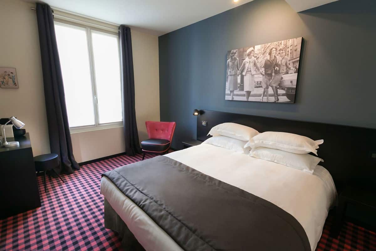 A hotel room with a large bed, a desk with a lamp, a chair, a window with curtains, and a wall-mounted picture.