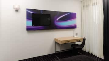 A hotel room with a flat-screen TV mounted on the wall, a desk with a chair, and a window with sheer curtains.