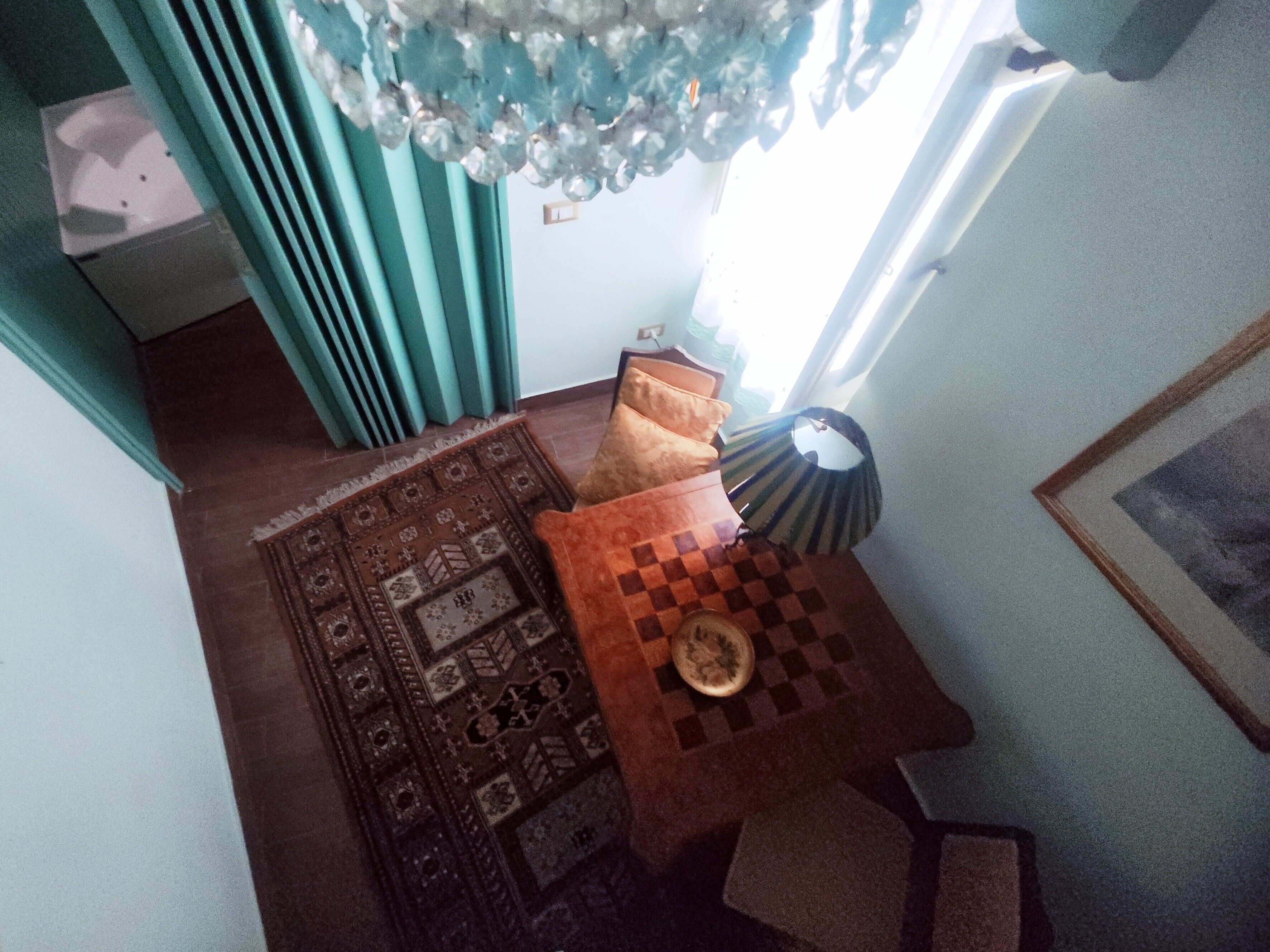 A room with a patterned rug, a small table with a chessboard, a lamp, and a window with curtains.