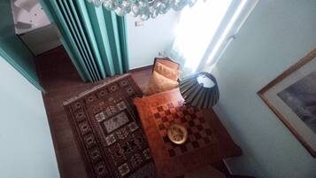 A room with a patterned rug, a small table with a chessboard, a lamp, and a window with curtains.