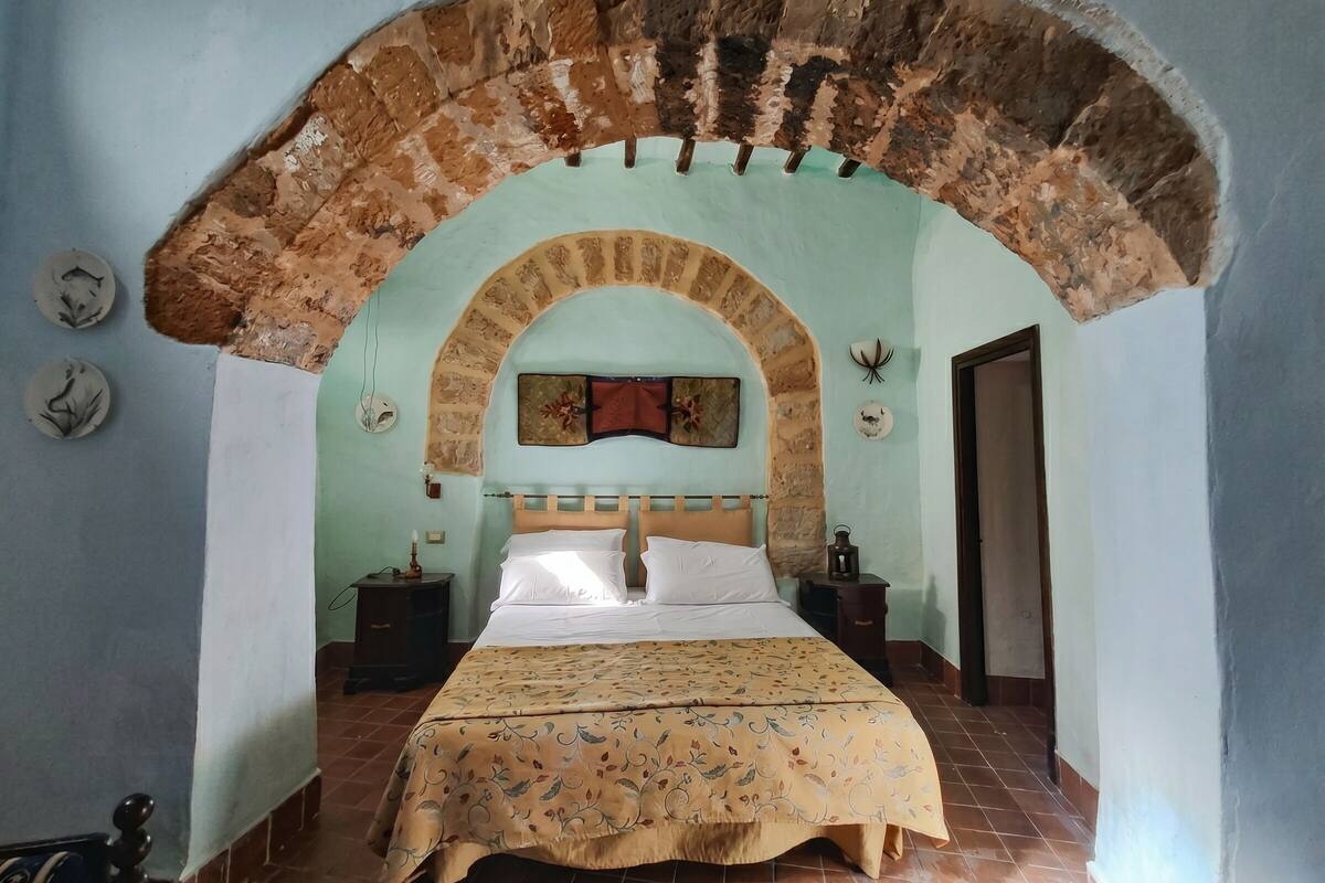 Een slaapkamer met een bed onder een gewelfde stenen constructie, een nachtkastje, een lamp en een decoratie aan de muur.