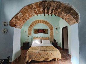 A bedroom with a bed under an arched stone structure, a nightstand, a lamp, and a wall-mounted decoration.
