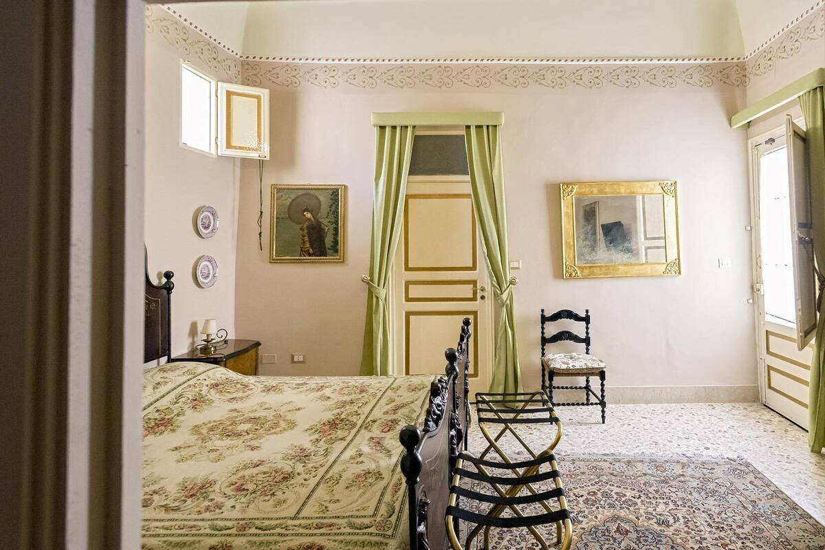 Een slaapkamer met een bed, een stoel, een spiegel en een deur met groene gordijnen.