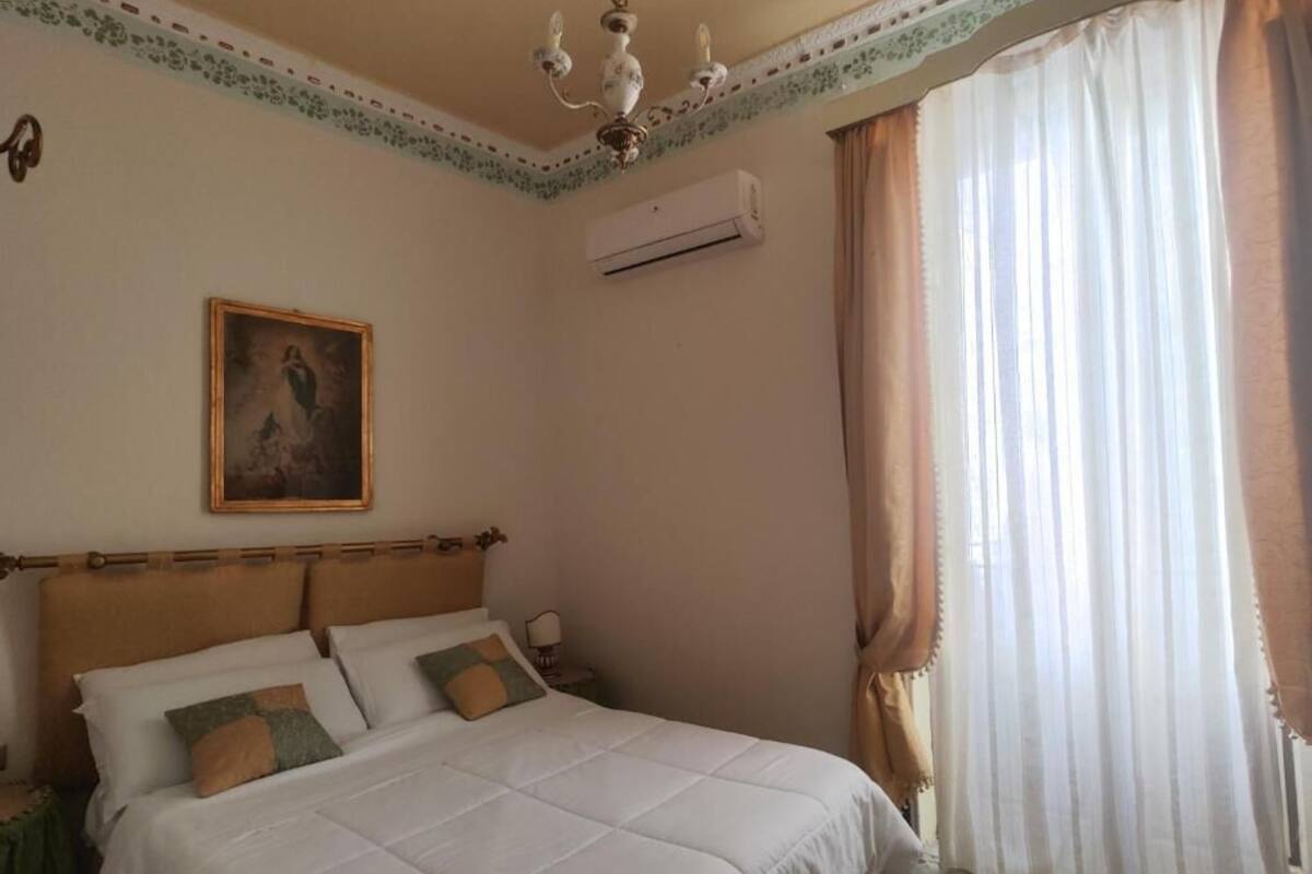 Een slaapkamer met een bed, een wandairconditioner, een kroonluchter en een raam met gordijnen.