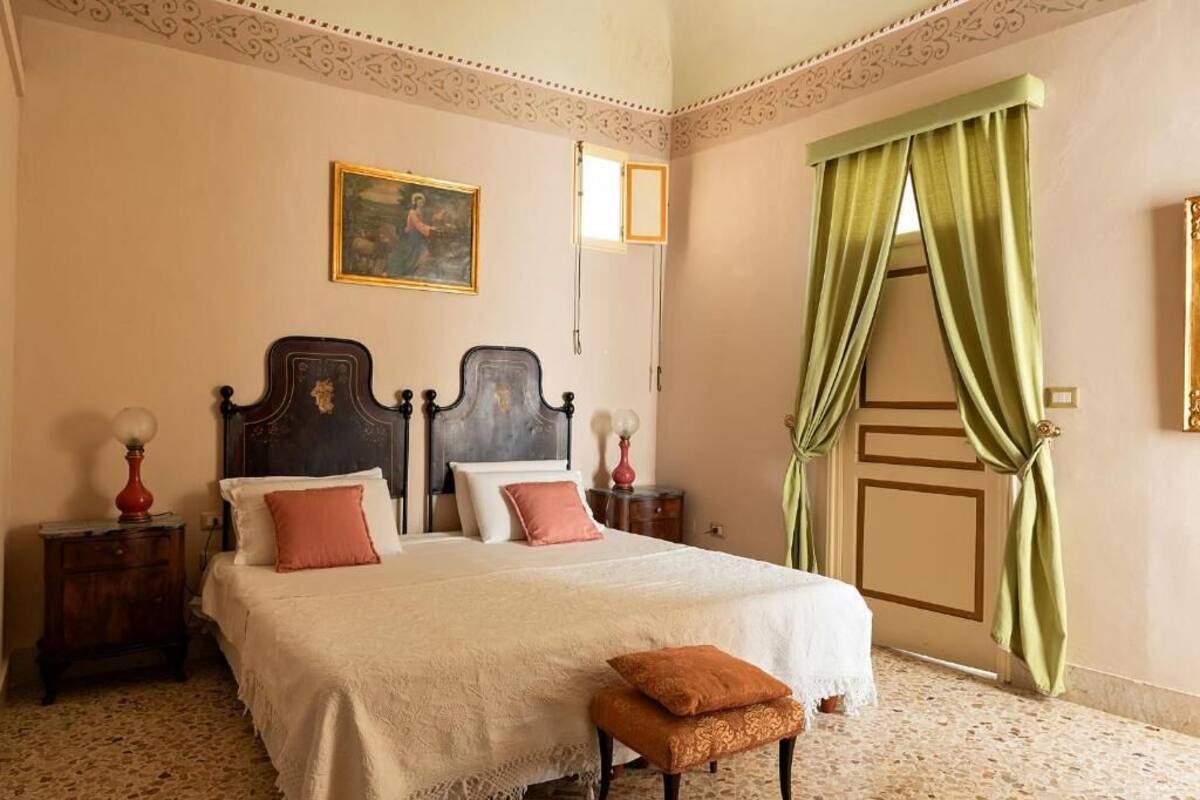 Een slaapkamer met een groot bed, twee nachtkastjes, een bankje, een schilderij aan de muur en een groen gordijn voor het raam.