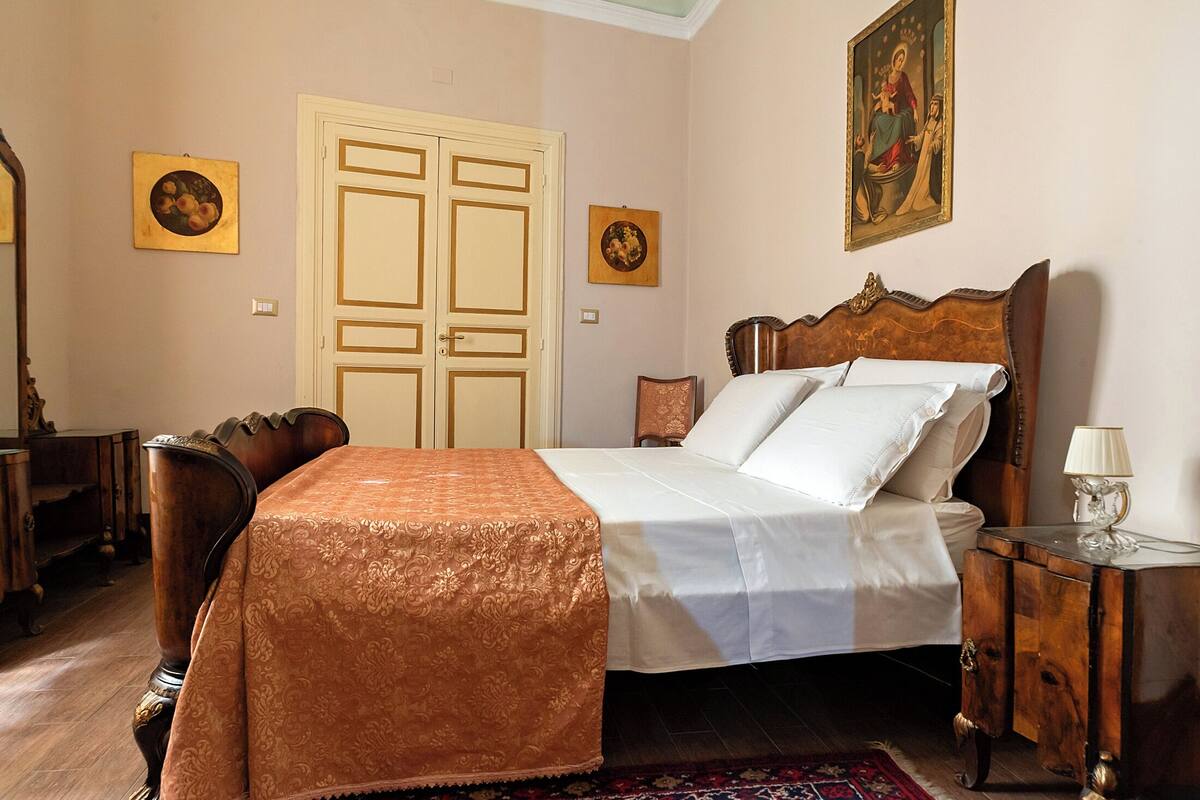 Een slaapkamer met een bed, een houten nachtkastje, een stoel, een deur en twee ingekaderde foto's aan de muur.