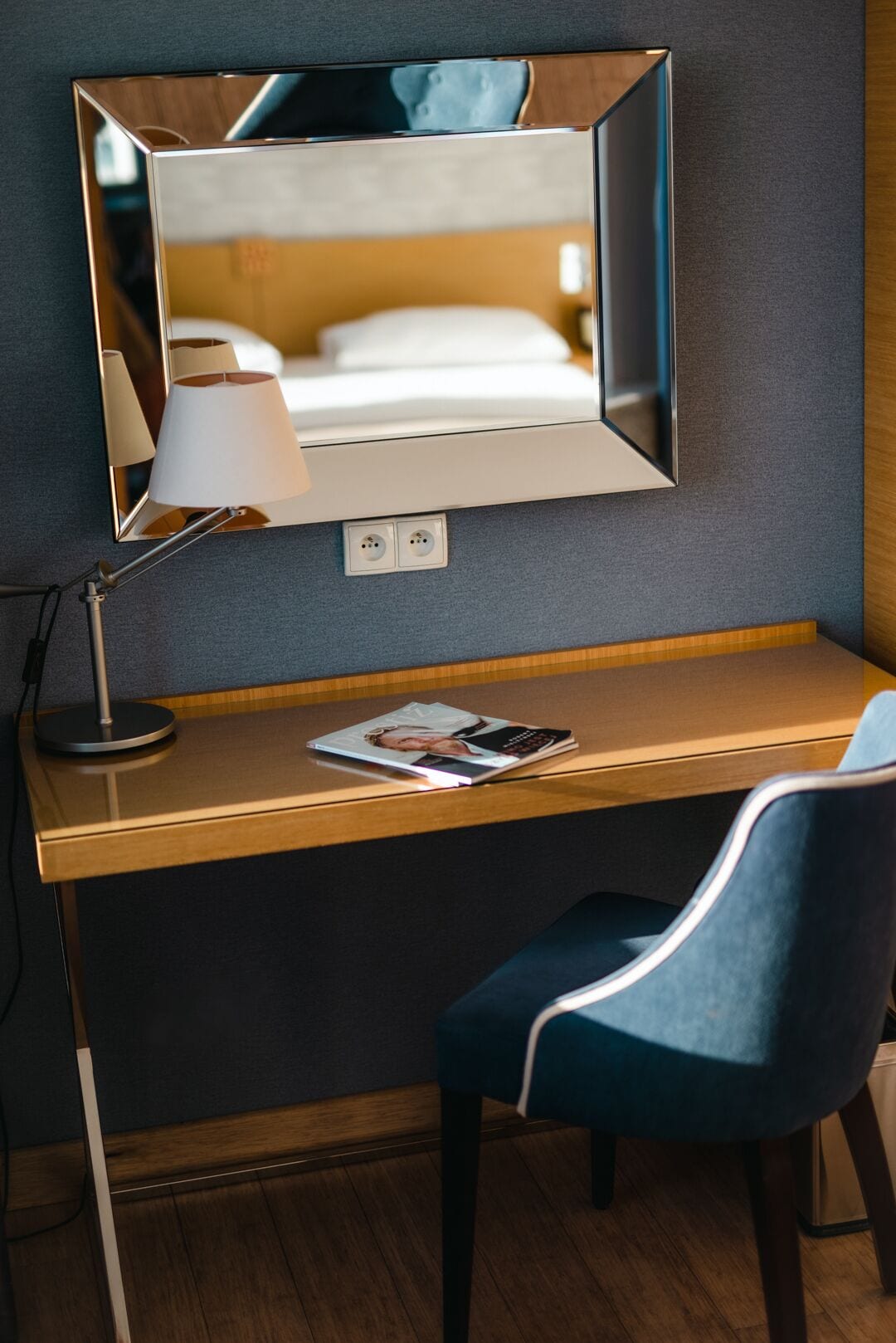Habitación de hotel con escritorio de madera, una silla azul, lámpara y espejo que refleja una cama.