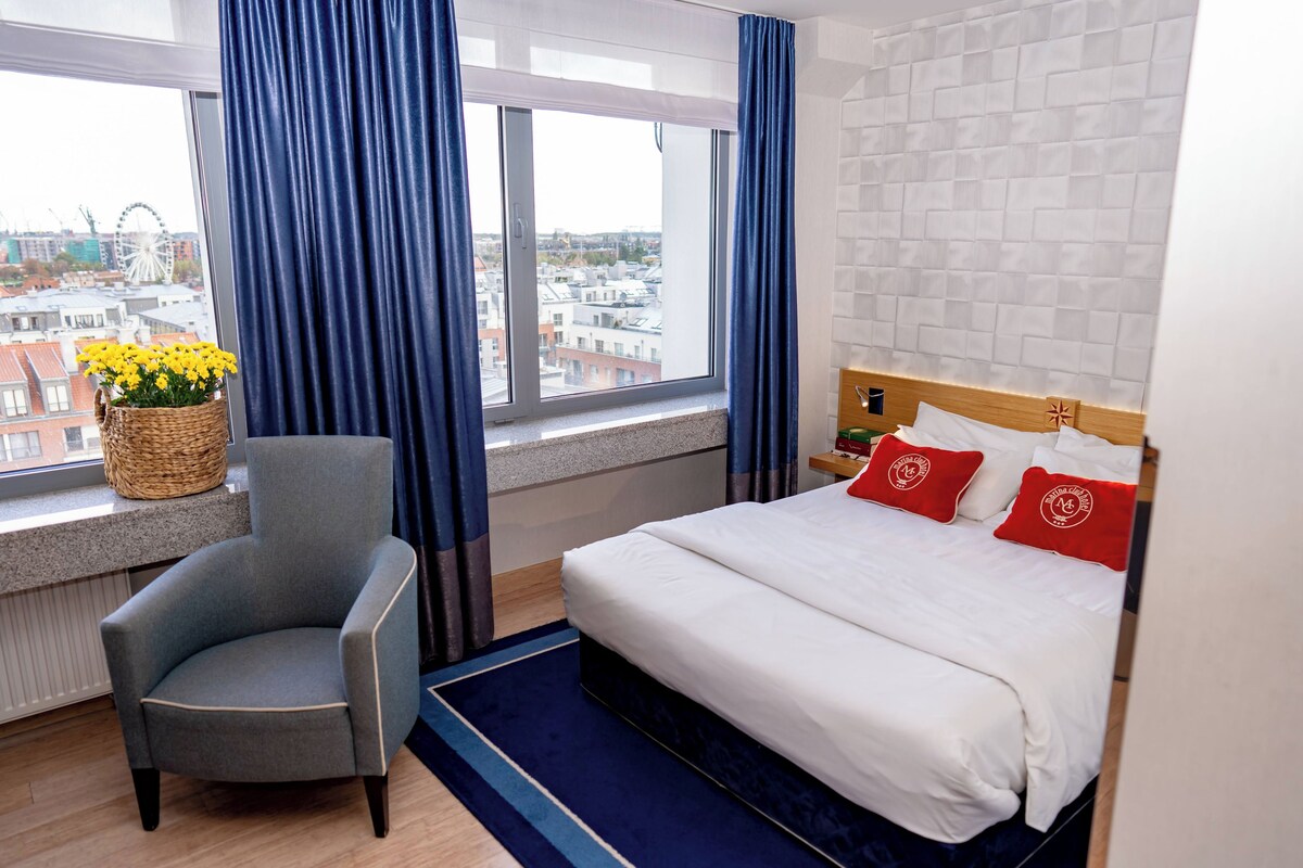 Habitación de hotel con cama, una silla, ventana con cortinas y vistas a la ciudad, además de un cesto con flores.