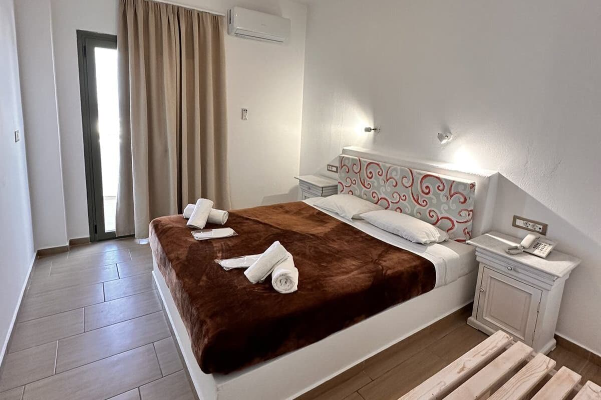 Une chambre d’hôtel comprenant un lit, une table de chevet avec un téléphone, une commode, un banc en bois, une porte et un appareil de climatisation.