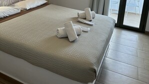 A hotel room with a large bed, a bedside table with a lamp, a view of the sea through the balcony doors, and an air conditioning unit on the wall.