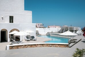 A pool area with lounge chairs and umbrellas, a stone wall, and a building with arched doorways.