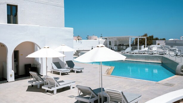 A rooftop pool area with white lounge chairs and umbrellas, a clear blue sky, and a view of the sea.