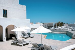 A rooftop pool area with white lounge chairs and umbrellas, a clear blue sky, and a view of the sea.