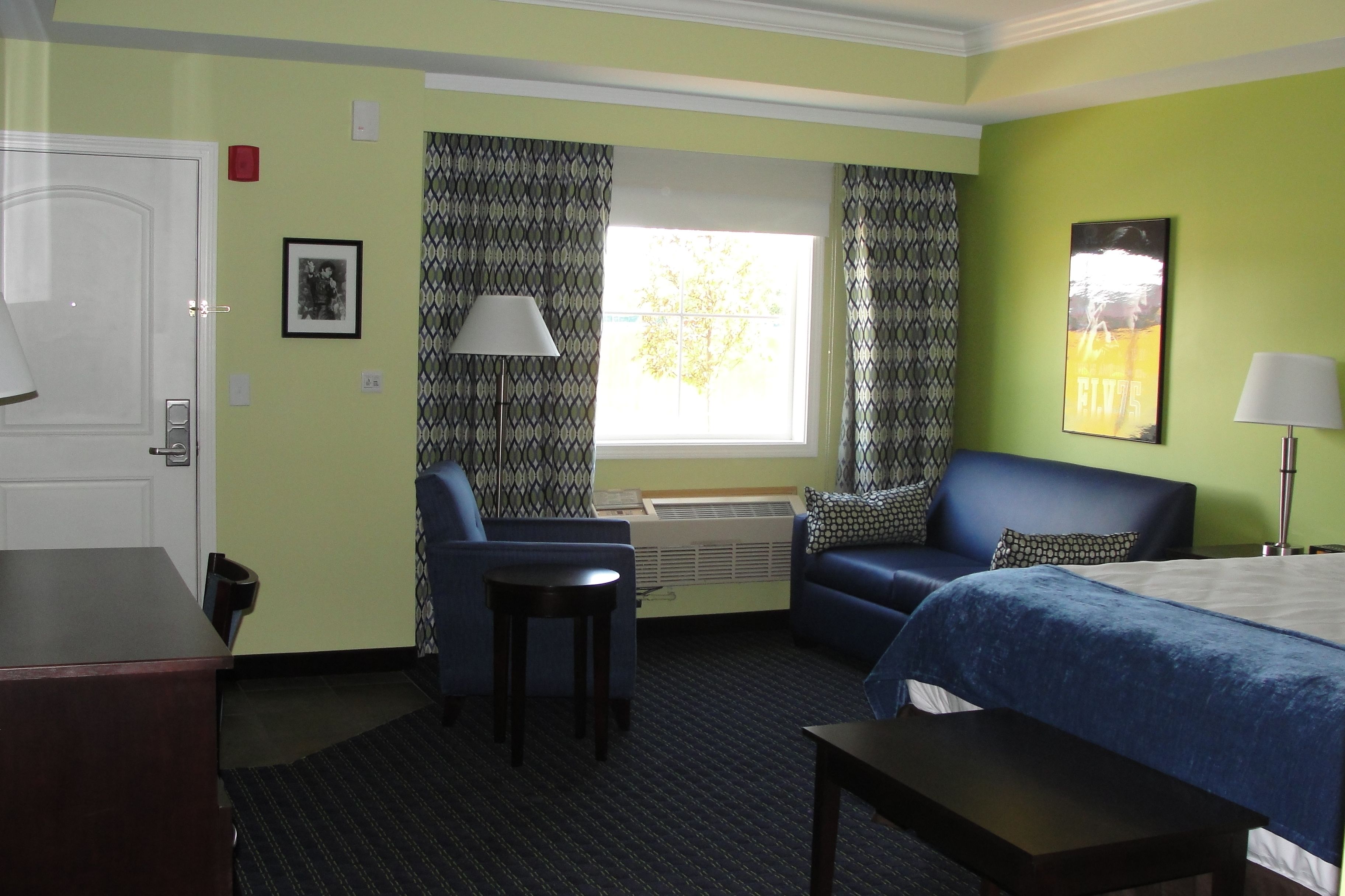 A hotel room with a bed, a desk, a chair, a small table, a window with curtains, a picture on the wall, and an air conditioning unit.