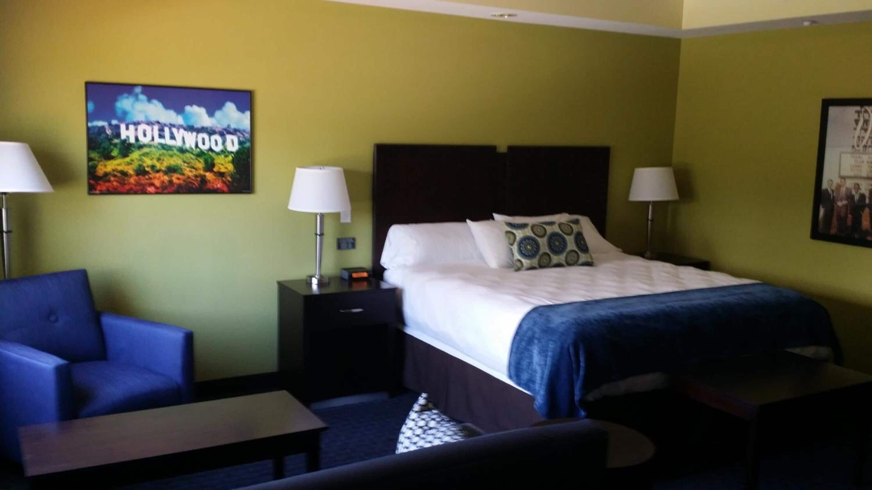 A hotel room with a bed, two nightstands, a blue armchair, a small table, and a picture of Hollywood on the wall.
