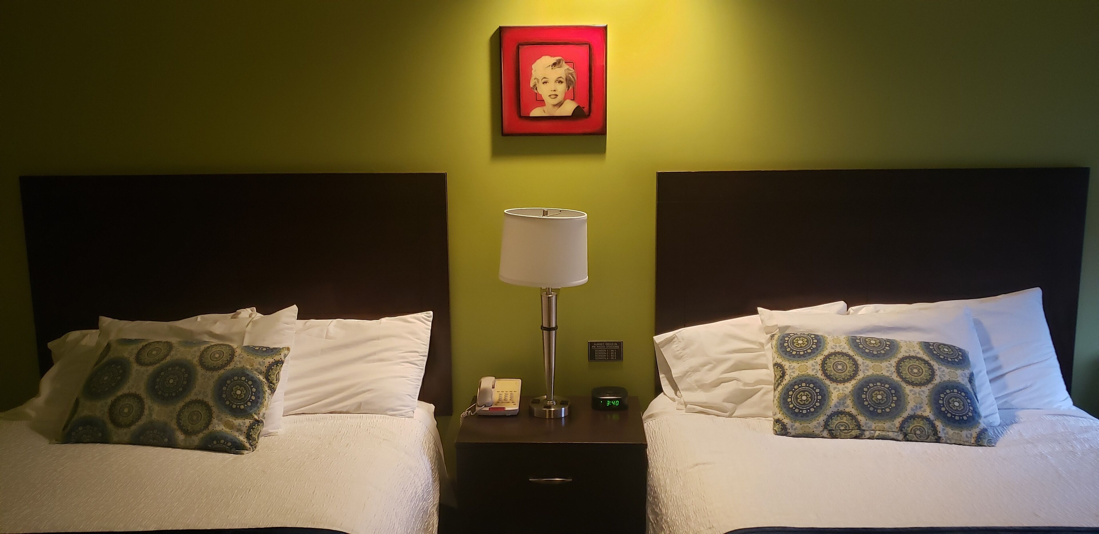 A hotel room with two beds, each with patterned pillows, a bedside lamp, a nightstand with a phone and clock, and a framed picture on the wall.