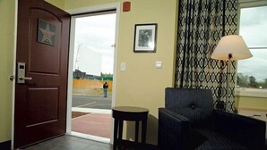 Un vestíbulo de hotel con una puerta marrón, una mesita redonda pequeña, un sillón azul, cortinas estampadas y un cuadro en la pared.