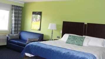 A hotel room with a bed, a blue sofa, a patterned curtain, a wall poster, and a nightstand with a lamp.