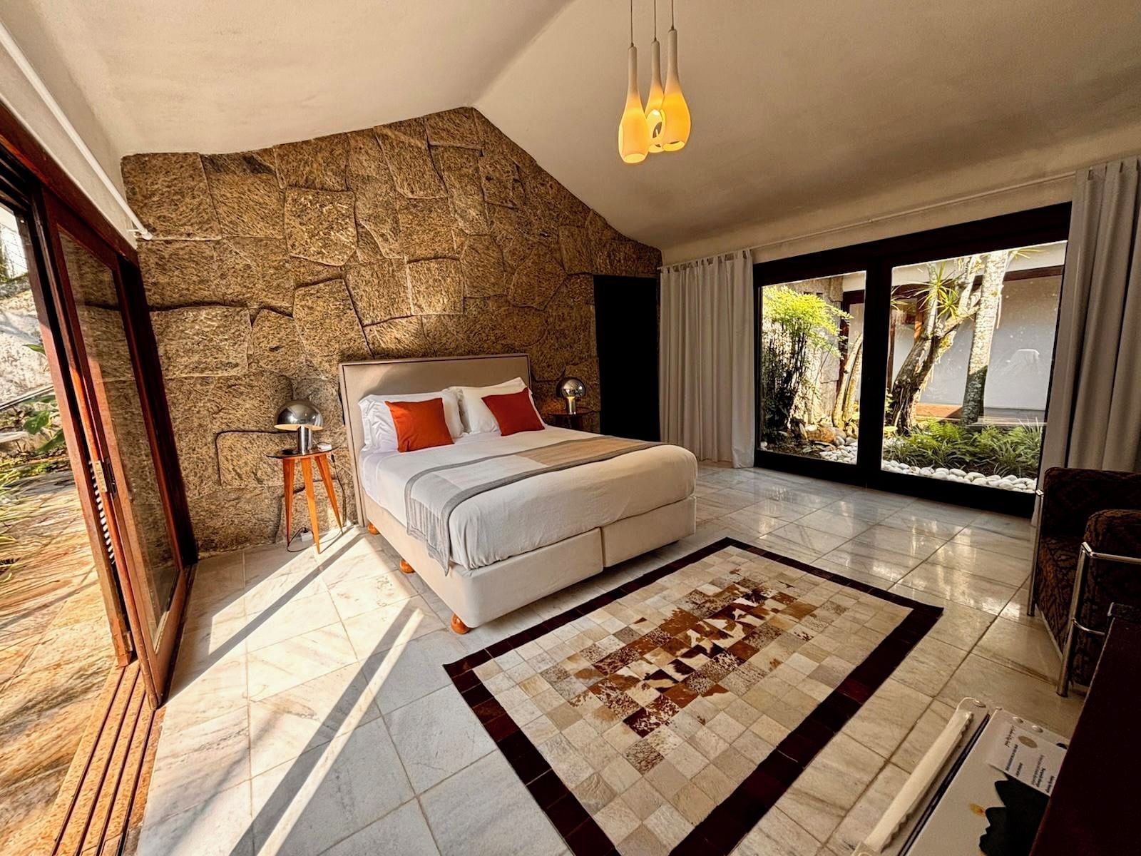 A modern bedroom with a stone accent wall, a large bed with pillows, a side table, and a view of greenery through a glass door.