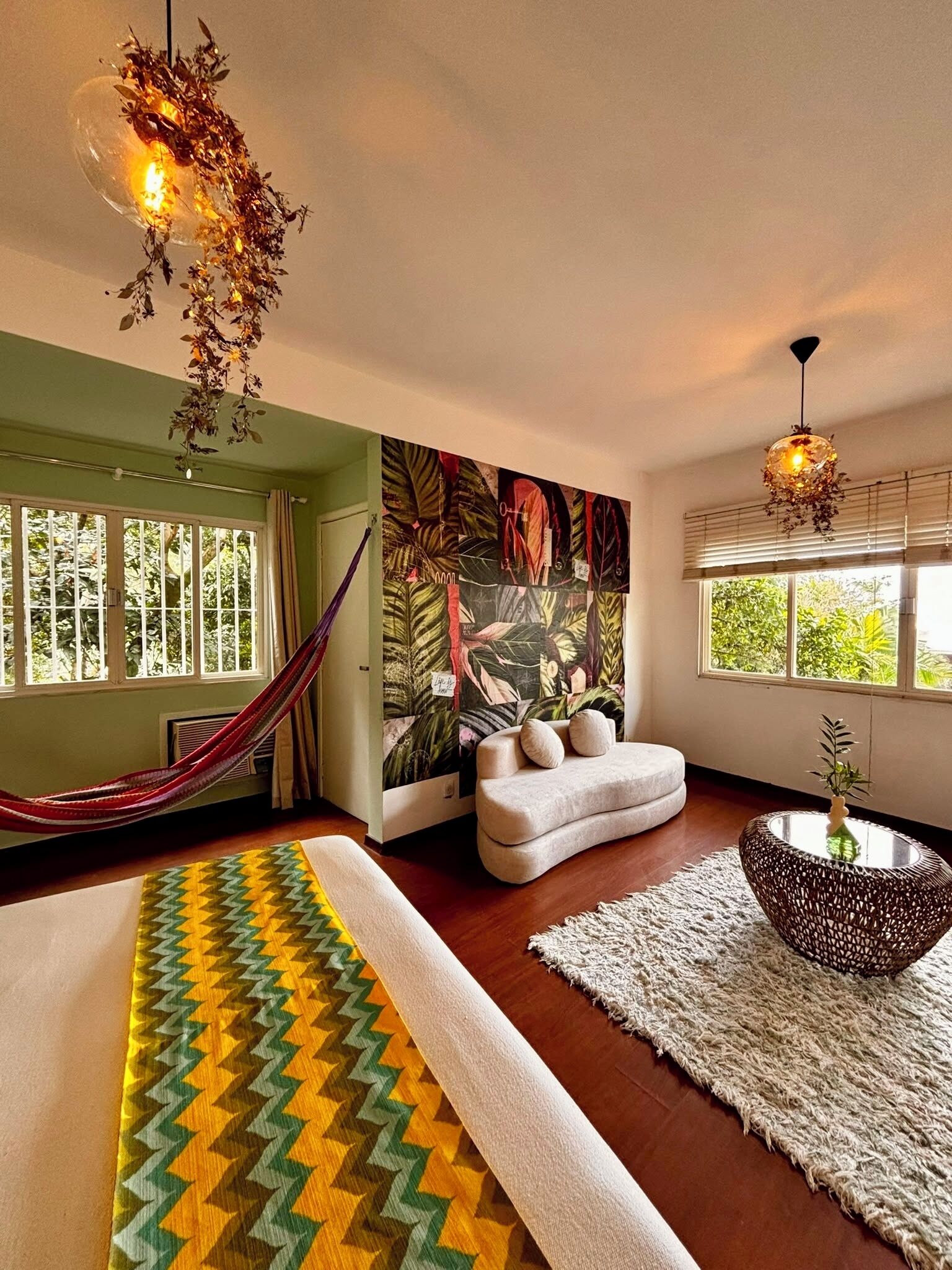 A room with a hammock, a large wall mural, a round table, and a view of greenery through the windows.