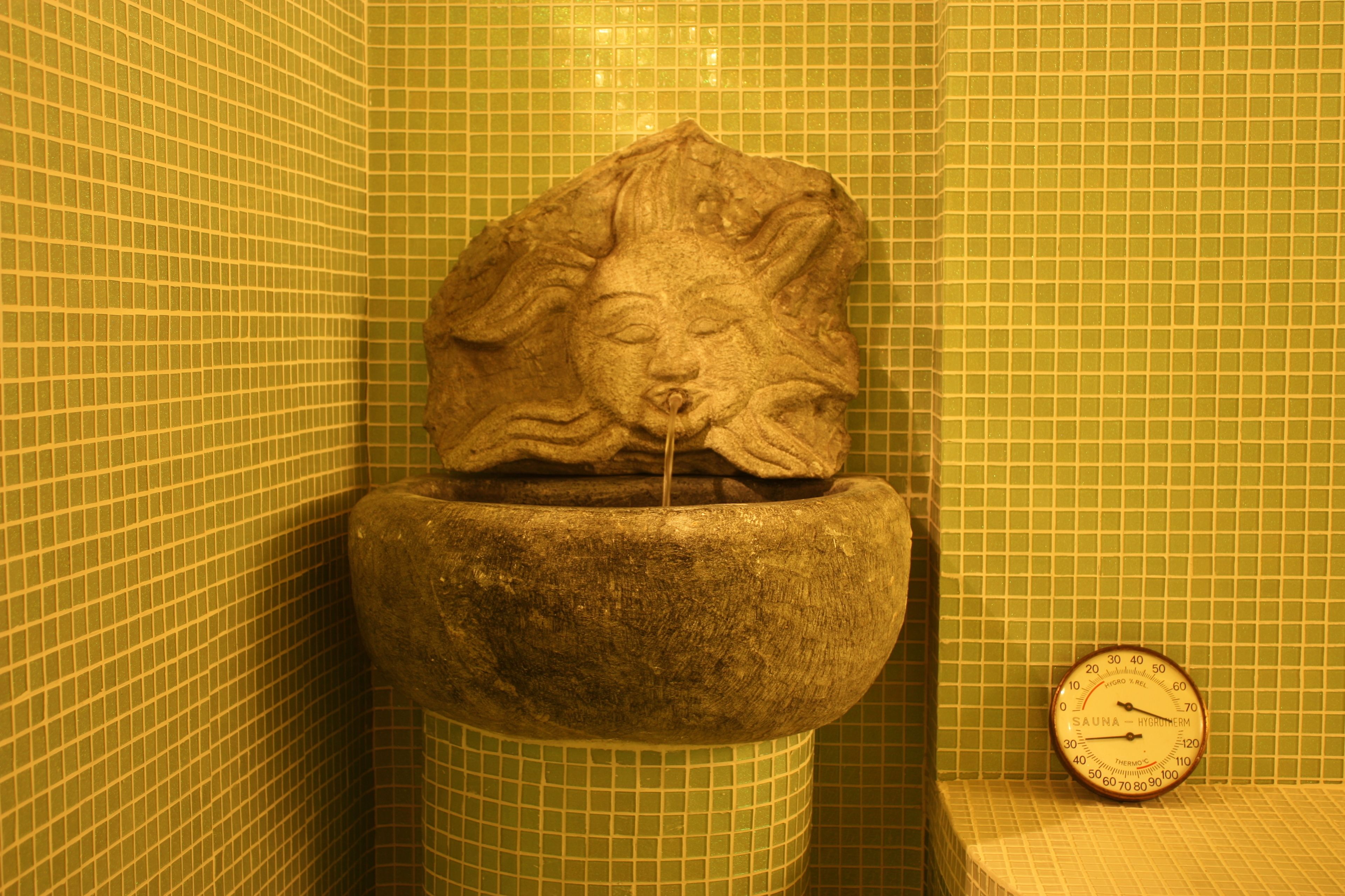 A stone fountain with a lion's head, a clock, and green tiled walls.