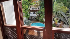 Vista a través de una ventana con piscina, rodeada de exuberante vegetación y un edificio blanco.