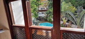 Vista a través de una ventana con piscina, rodeada de exuberante vegetación y un edificio blanco.