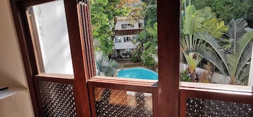 Vista a través de una ventana con piscina, rodeada de exuberante vegetación y un edificio blanco.