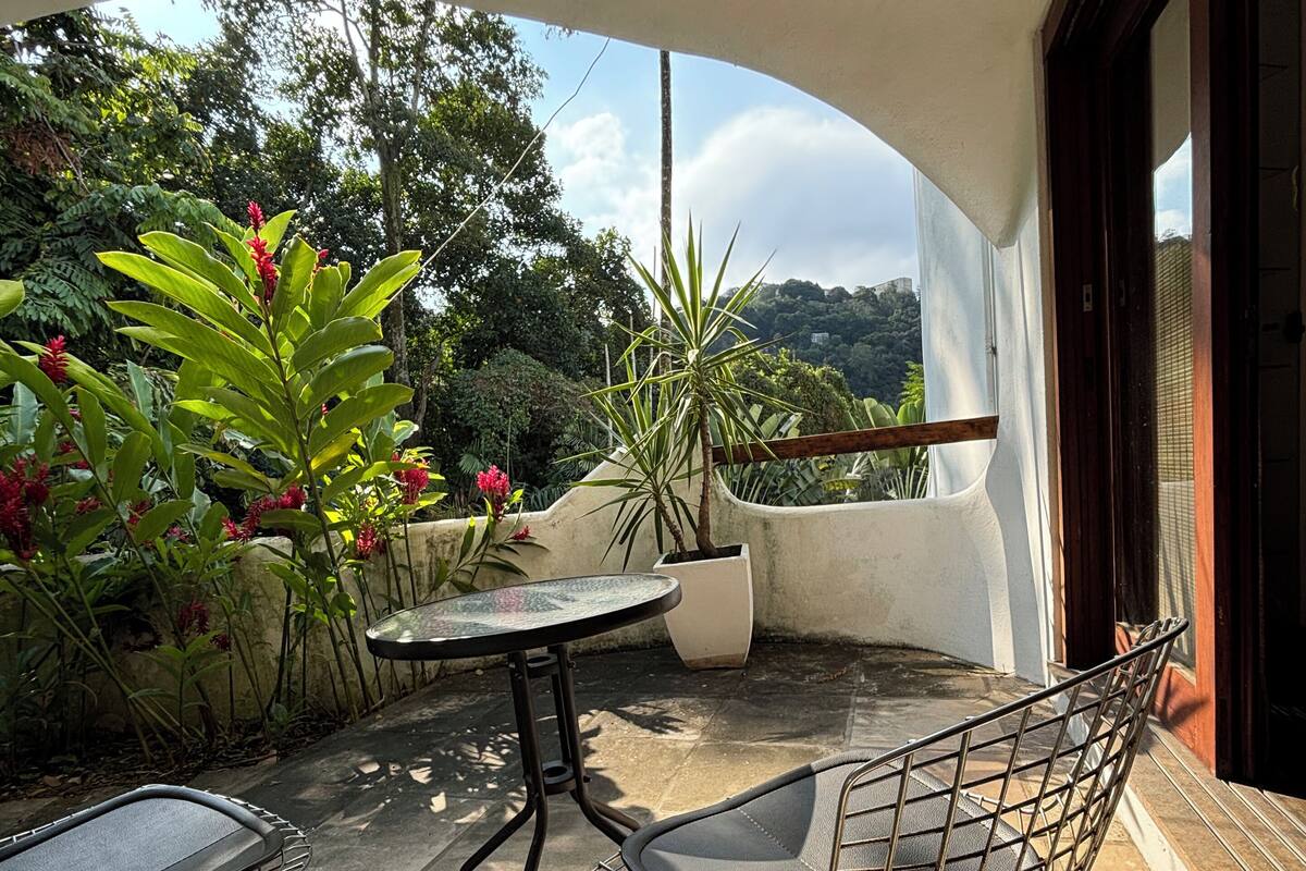 Un patio al aire libre con una mesa redonda y dos sillas, rodeado de macetas con plantas y follaje.