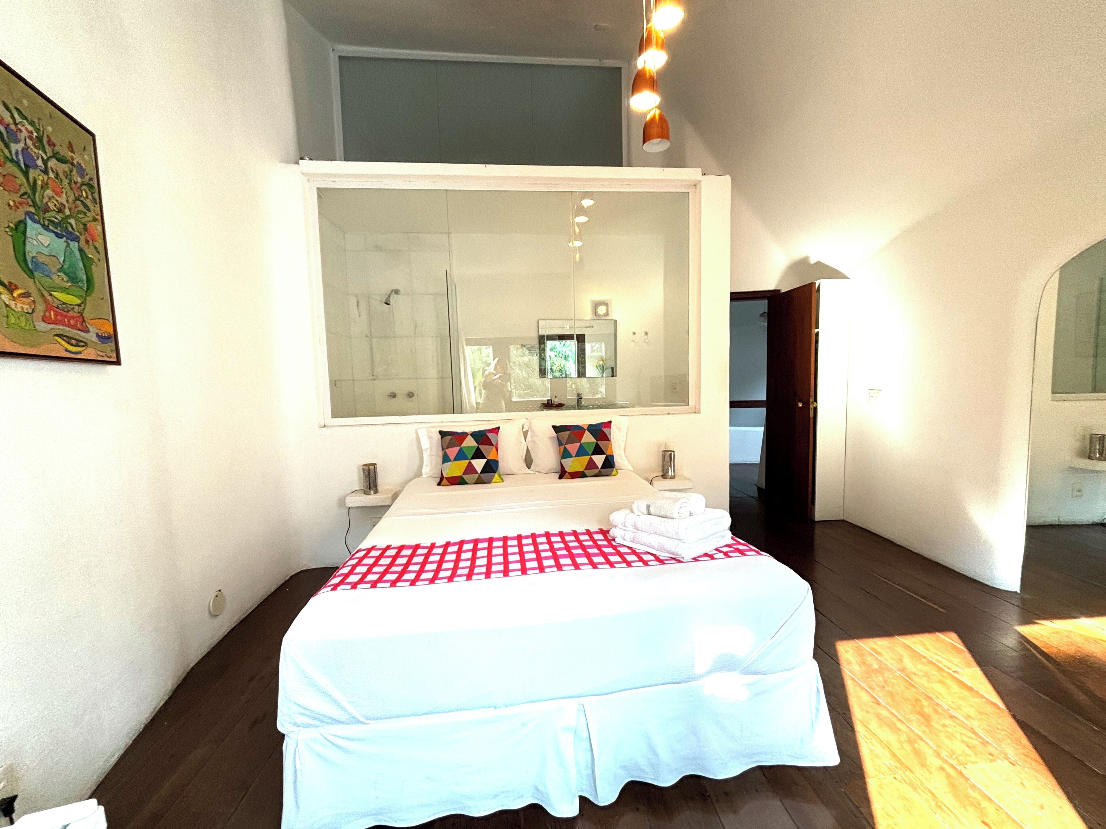 A bedroom with a large mirror, a bed with a checkered bedspread, two pillows, a nightstand, and a wooden floor.