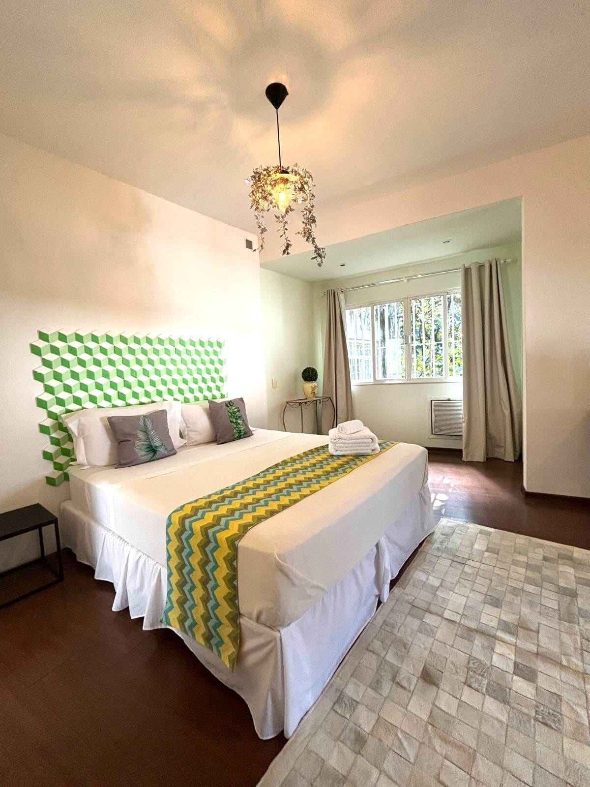 Un dormitorio con una cama, una mesita de noche, una lámpara colgante, una ventana con cortinas y vistas al exterior.