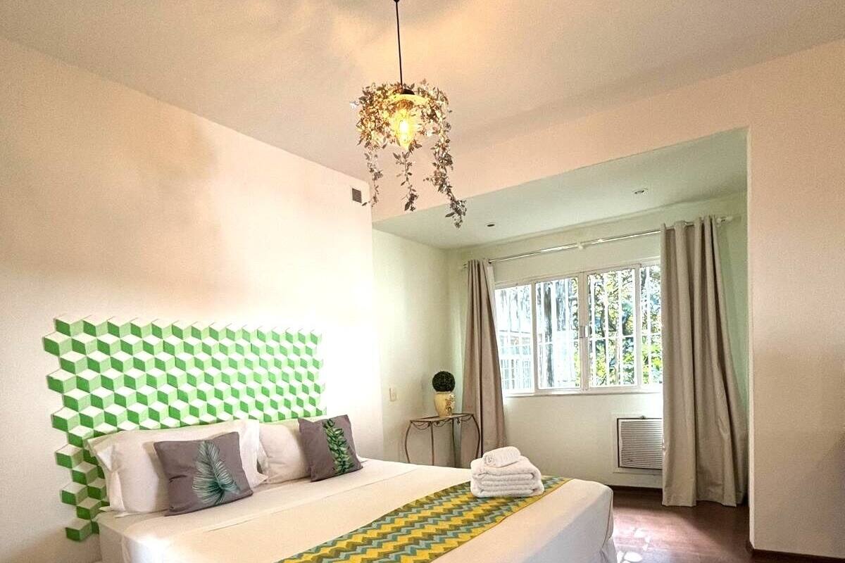 Un dormitorio con una cama, una mesita de noche, una lámpara colgante, una ventana con cortinas y vistas al exterior.