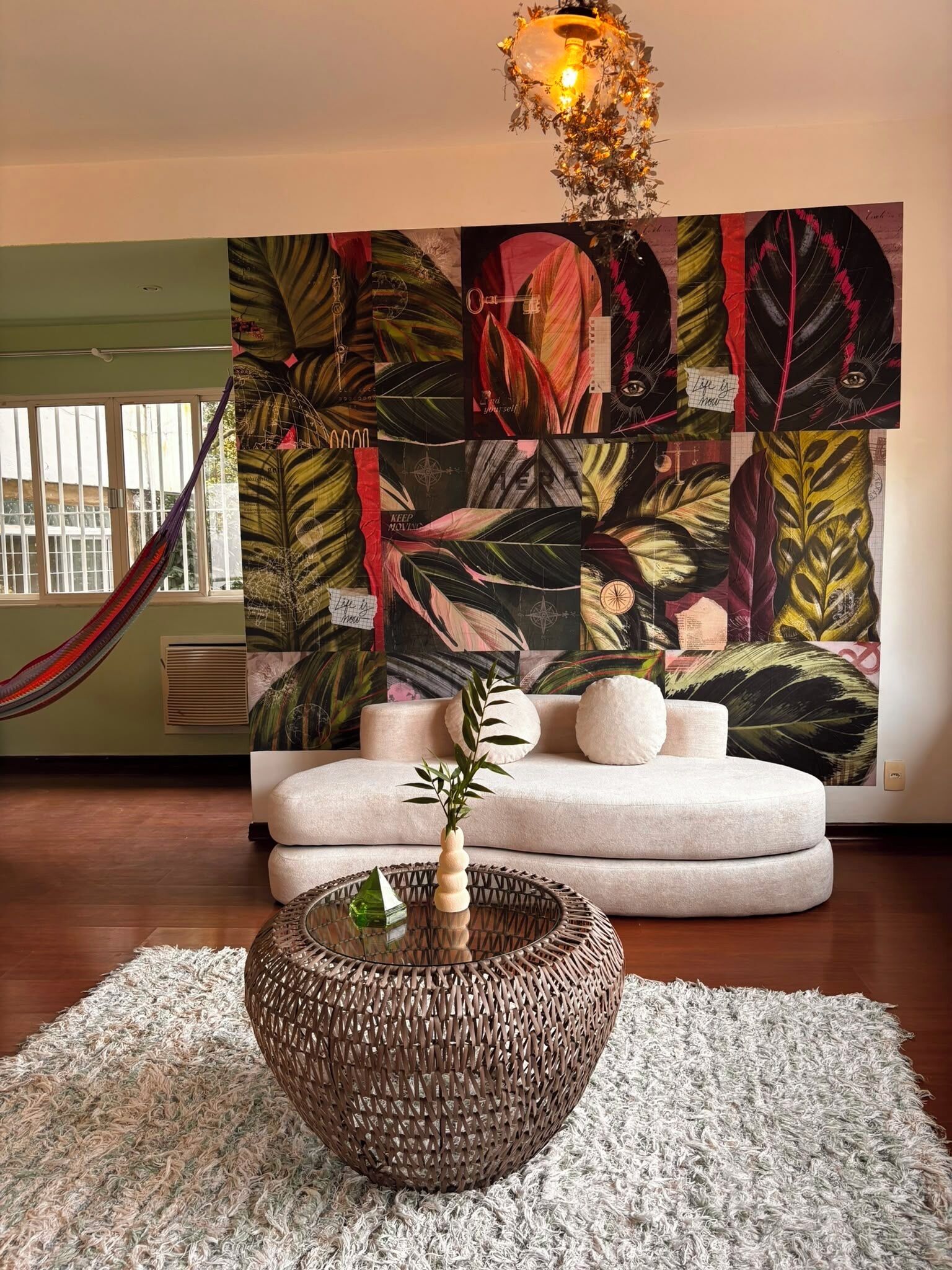 Una habitación con un cuadro grande de hojas tropicales, una mesa redonda tejida y una hamaca.