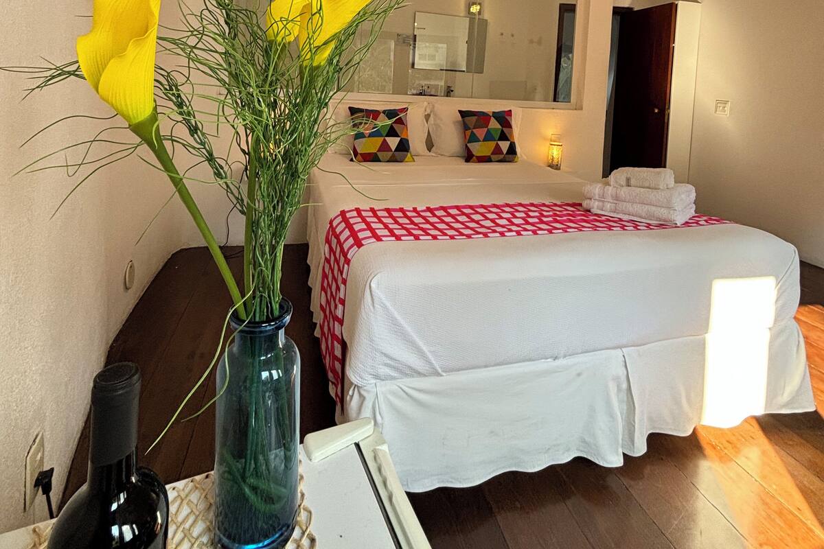 Un dormitorio con una cama, un espejo, un jarrón con flores y una botella sobre una mesa.
