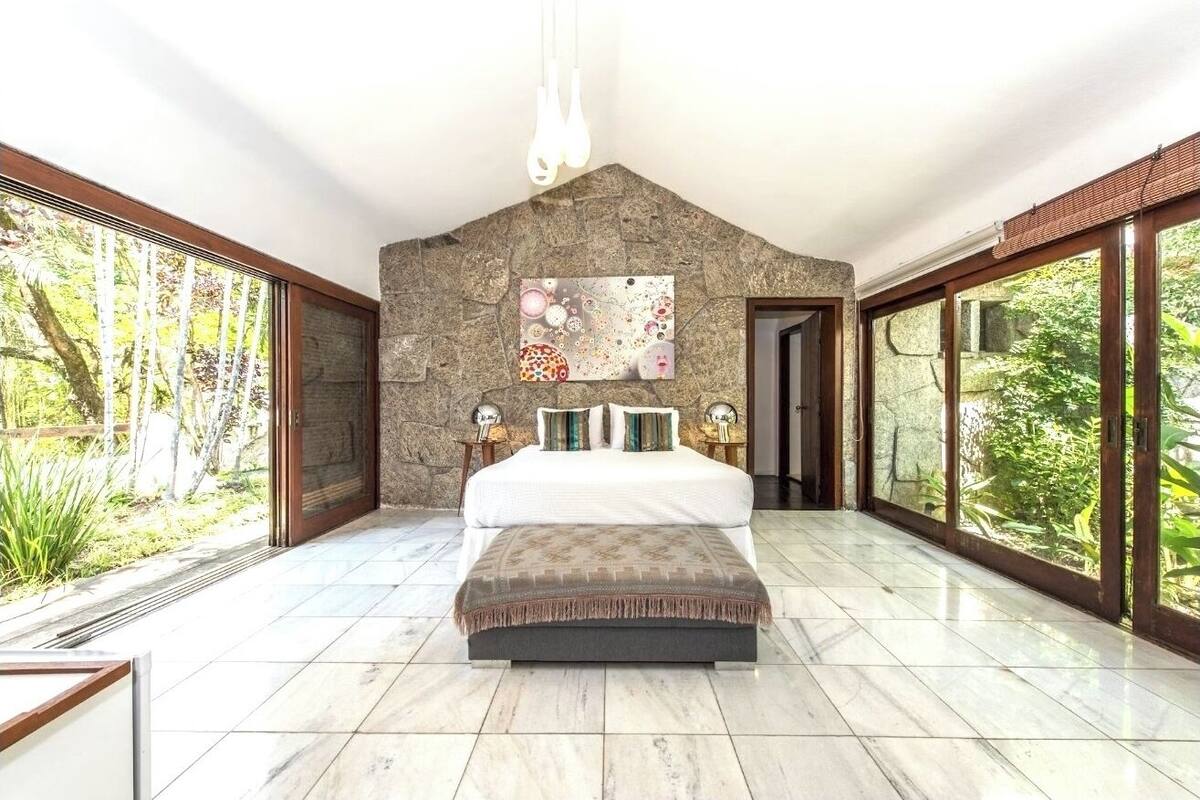 Un dormitorio con pared de piedra, una cama con almohadas, un cuadro y ventanas grandes.