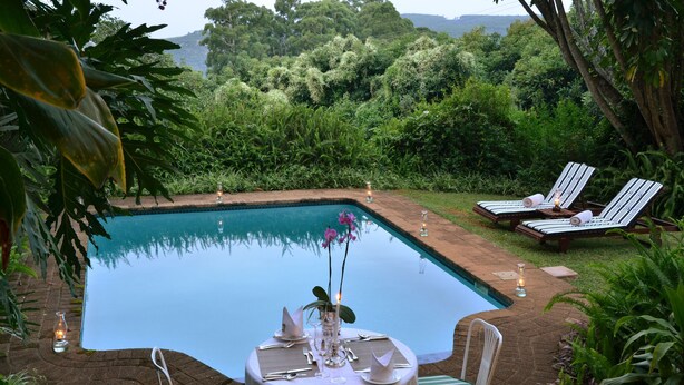 A poolside dining area with a table set for two, surrounded by lush greenery and overlooking a pool. - Highgrove House (Mbombela)