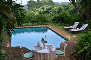 A poolside dining area with a table set for two, surrounded by lush greenery and overlooking a pool. - Highgrove House (Mbombela)