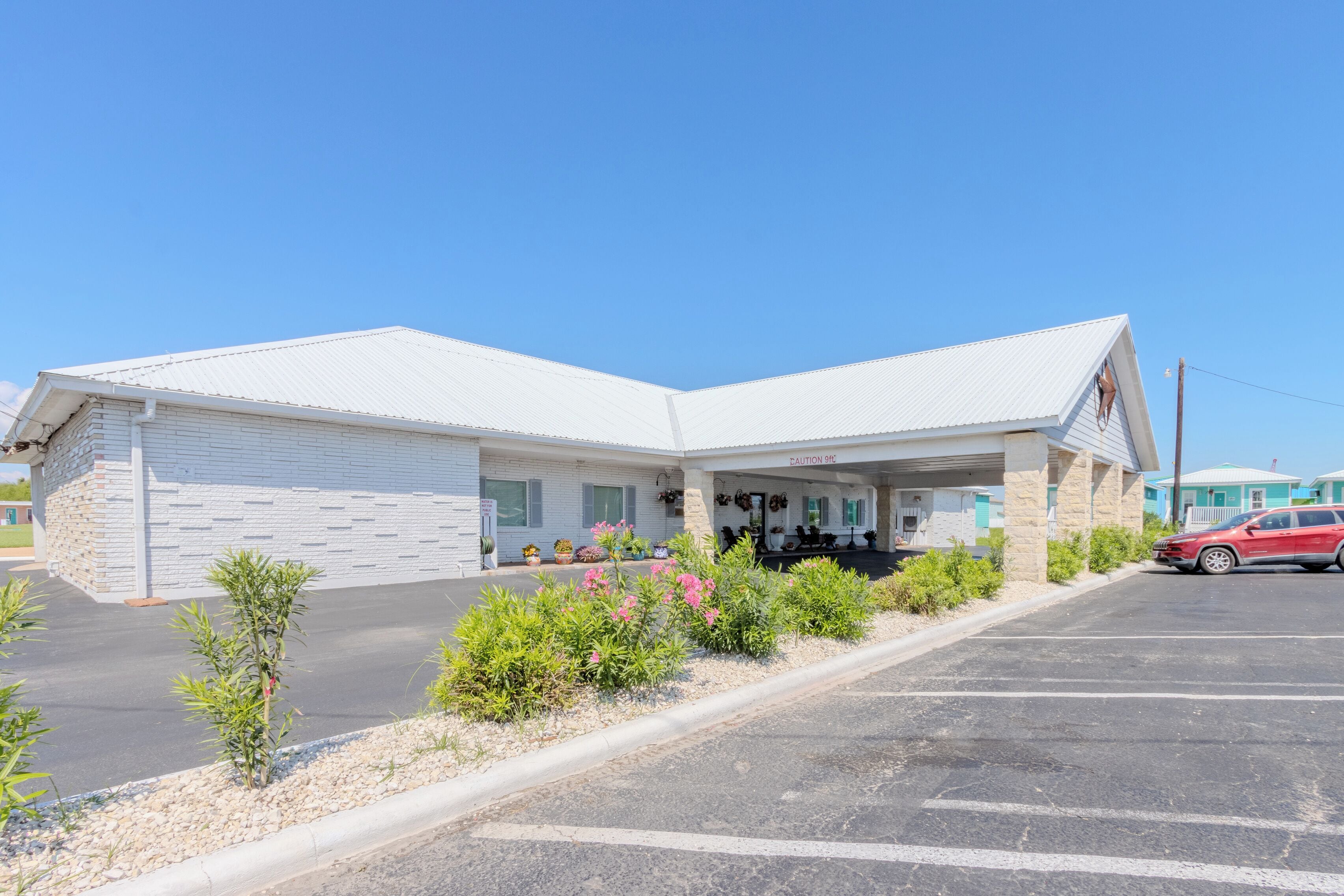 Un motel à l’extérieur blanc, doté d’un espace de stationnement couvert et où une voiture rouge est garée sur le parking.