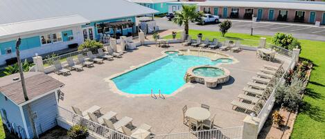 A swimming pool with a hot tub, surrounded by lounge chairs and tables, with a parking lot and buildings in the background.