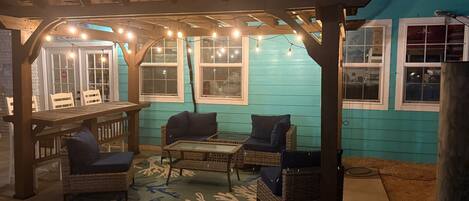 Un patio exterior con pérgola de madera, luces colgantes, mobiliario de mimbre y una mesa de vidrio.