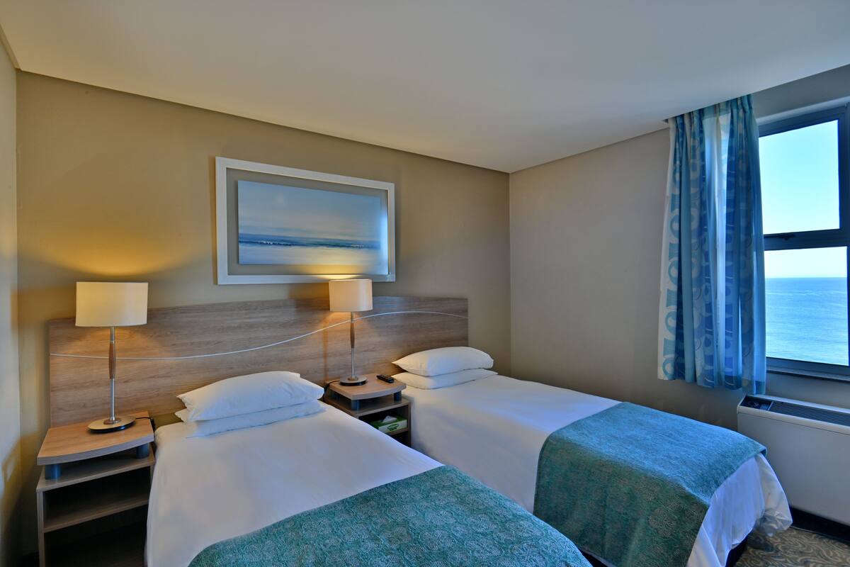 Habitación de hotel con dos camas, una mesita de noche con lámpara, un cuadro enmarcado en la pared y una ventana con cortinas.