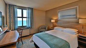 A hotel room with a bed, a desk, a chair, a window with curtains, a lamp, and a painting on the wall.