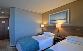 Una habitación de hotel con dos camas, un cuadro de una playa, una mesita de noche con lámpara y una puerta con cerradura de tarjeta.