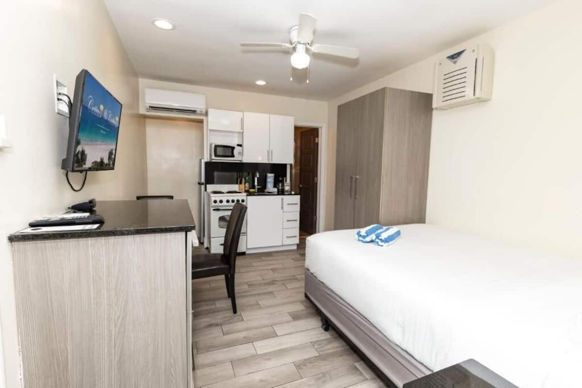 A compact hotel room with a kitchenette, a bed, a desk, and a ceiling fan.