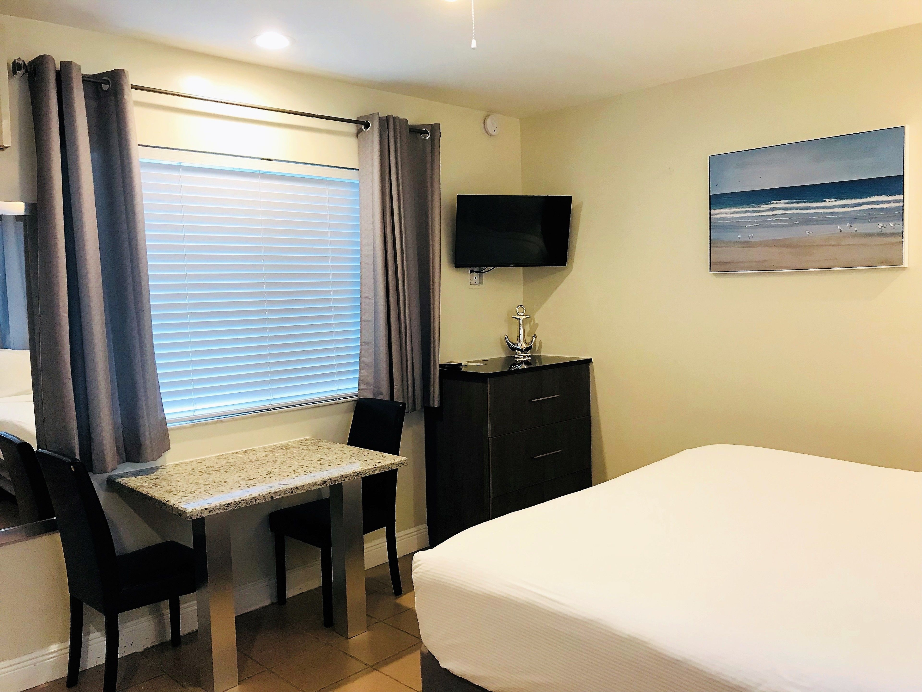 A hotel room with a bed, a desk with a chair, a TV, and a painting of a beach scene.