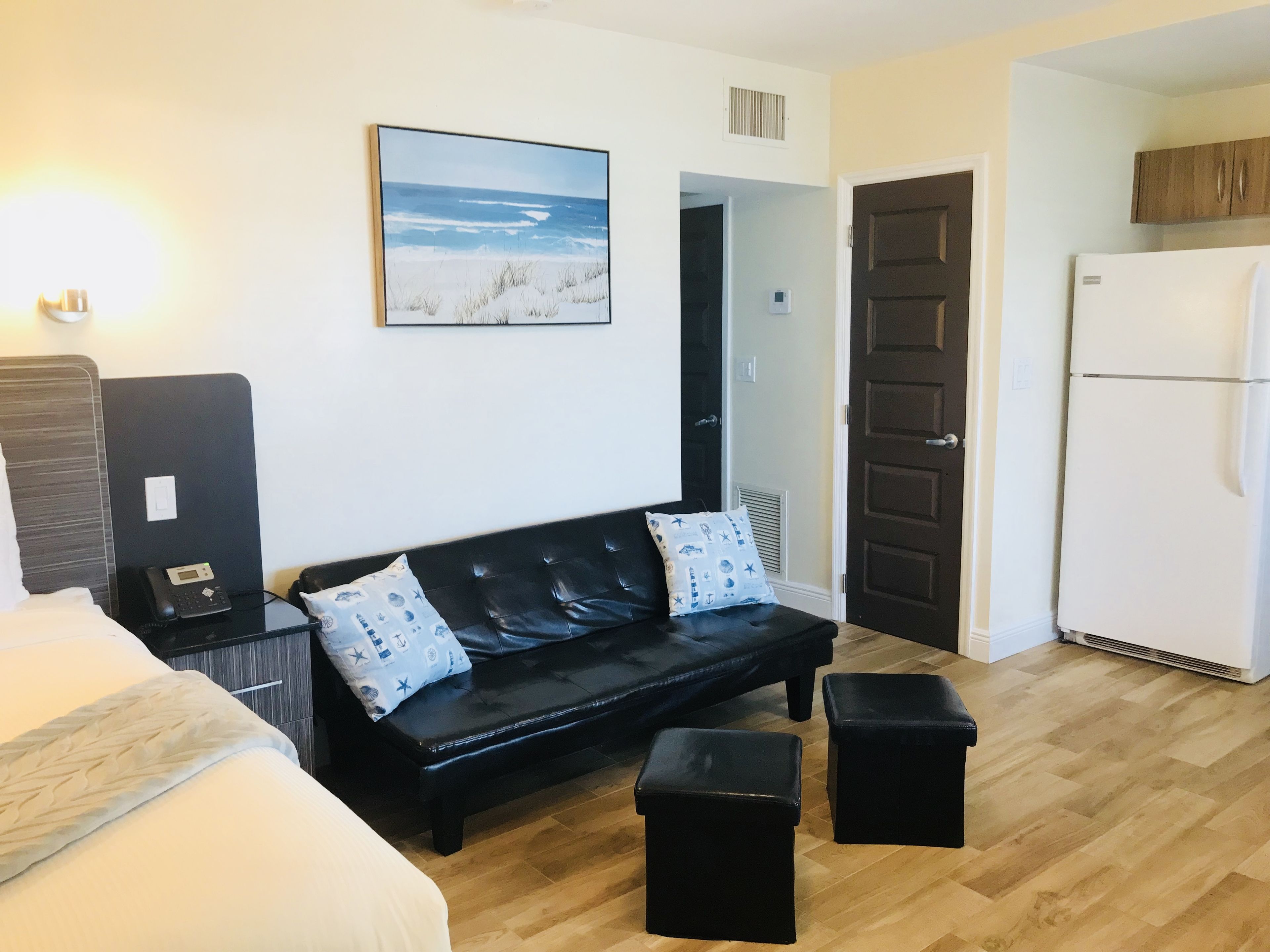 Una habitación con una cama, un sofá de cuero negro con cojines estampados, un pequeño banquito negro, un refrigerador blanco y una imagen colgada en la pared de una playa.