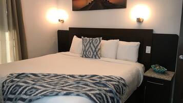 A neatly made bed with a patterned throw, a nightstand with a decorative bowl, and a sunset painting above the bed.