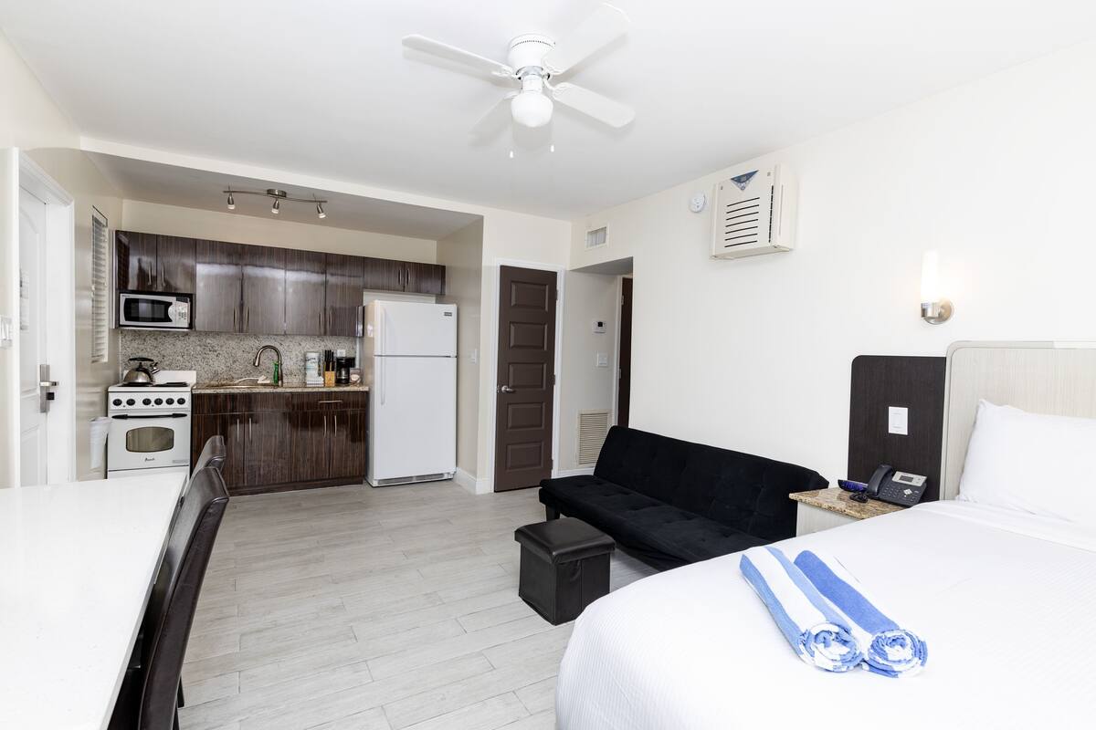 A hotel room with a kitchenette, a bed with white bedding, a black sofa, and a ceiling fan.